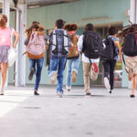 Group of school children wearing backpacks running down a covered outdoor school hallway toward a building entrance, captured from behind during the school day.