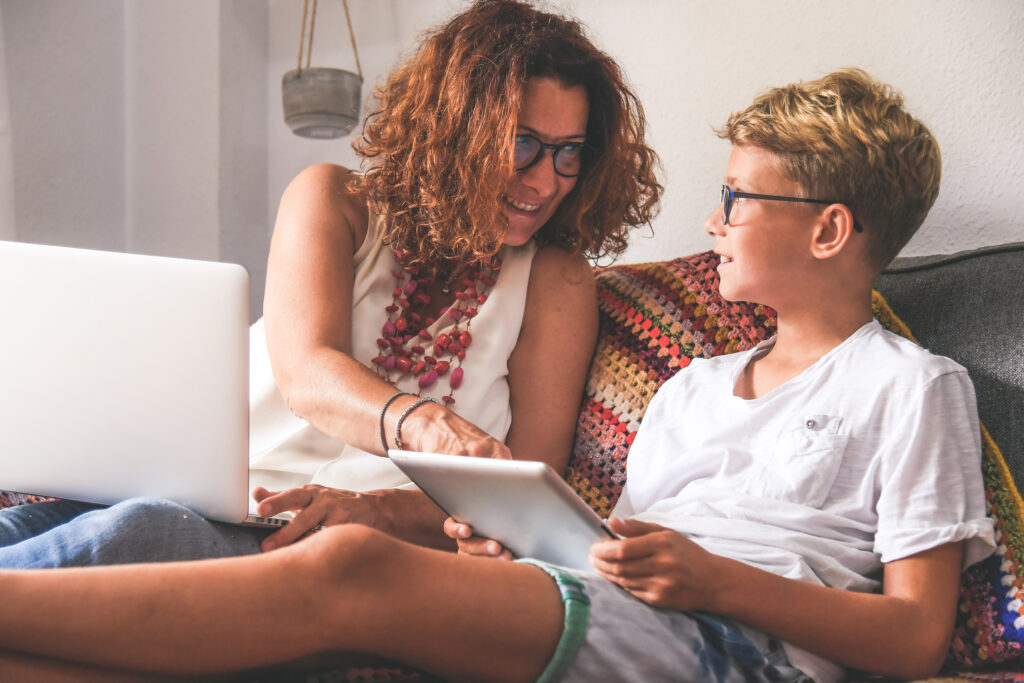 Mother and son sitting on a couch at home, smiling and interacting while using a tablet and laptop; casual indoor family moment with technology, homeschooling or online learning concept, both wearing glasses, relaxed lifestyle setting with cozy decor.