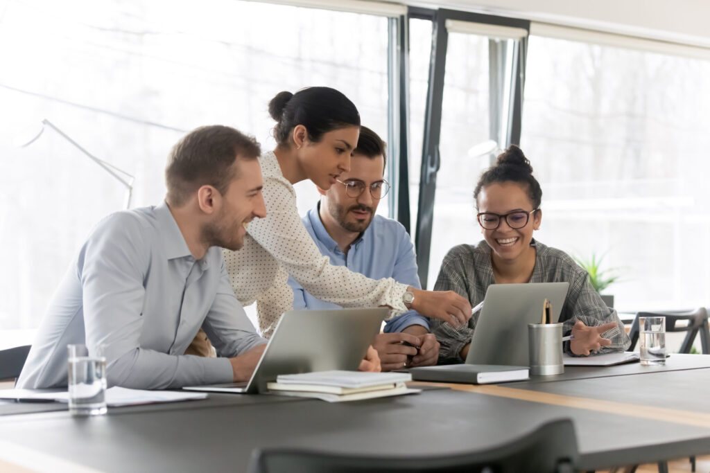 Smiling diverse young colleagues gather at desk in office look at laptop