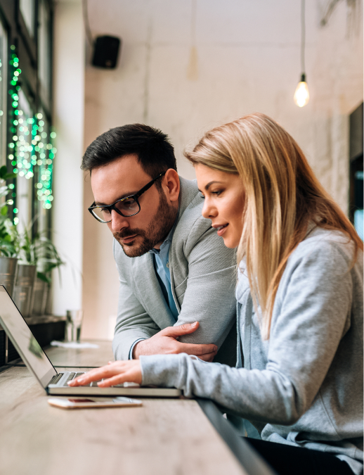 Two coworkers leaning over a laptop while discussing information together in a modern office or café workspace.