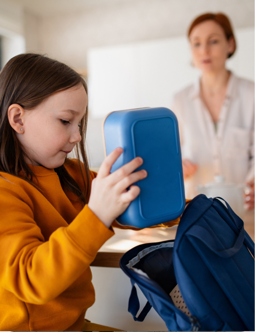 Young girl placing a blue lunch container into a backpack while an adult looks on in a home kitchen or dining area.