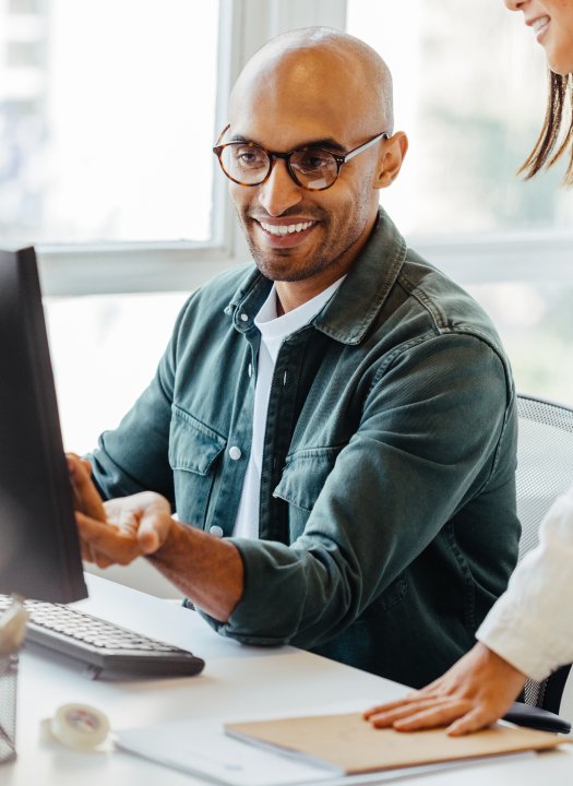 Smiling man wearing glasses working at a desktop computer while a coworker stands beside him in a bright office.