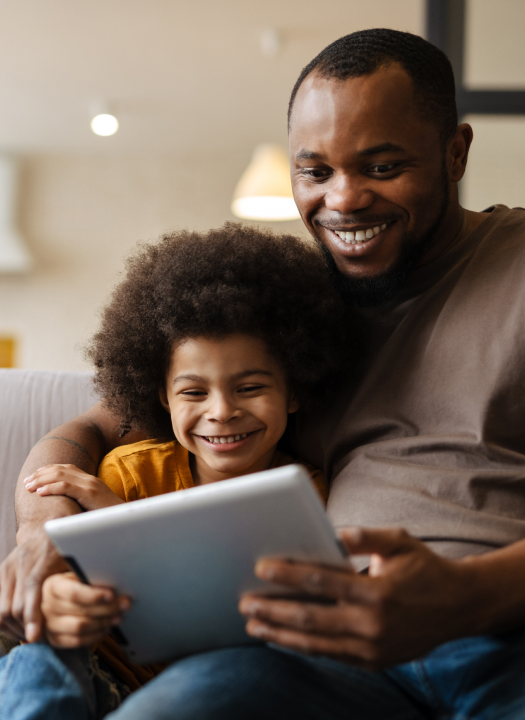 Smiling father and young child sitting together on a couch while looking at a tablet at home.