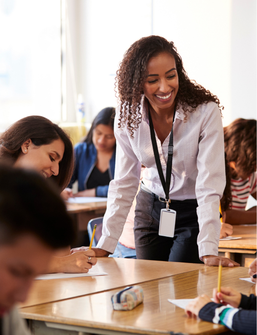Smiling teacher wearing a lanyard leaning over student desks while assisting students during a classroom activity.