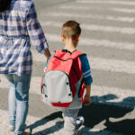 Adult holding a young child’s hand while walking with a school-age boy wearing a red backpack across a crosswalk outdoors.