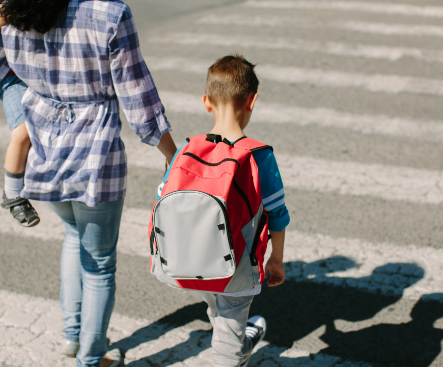 Adult holding a young child’s hand while walking with a school-age boy wearing a red backpack across a crosswalk outdoors.