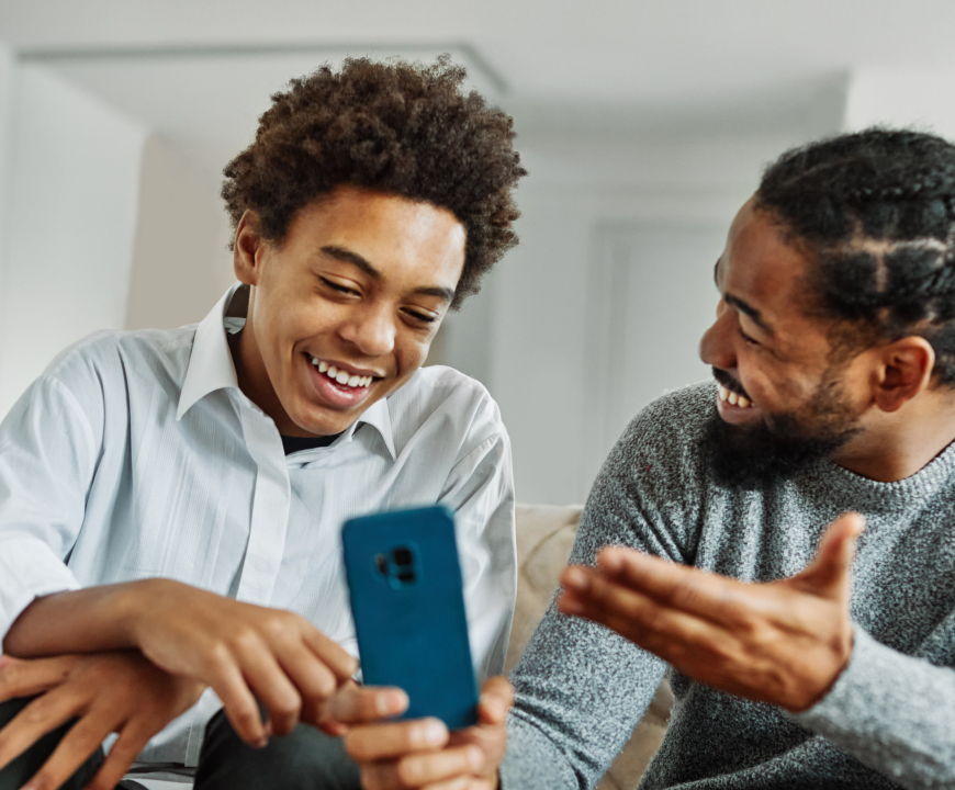 Teen boy and parent smiling while looking at a smartphone together during a casual conversation in a home setting.