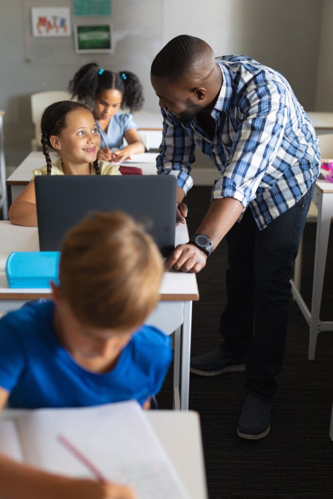 smiling african american young male teacher looking at caucasian elementary schoolgirl with laptop